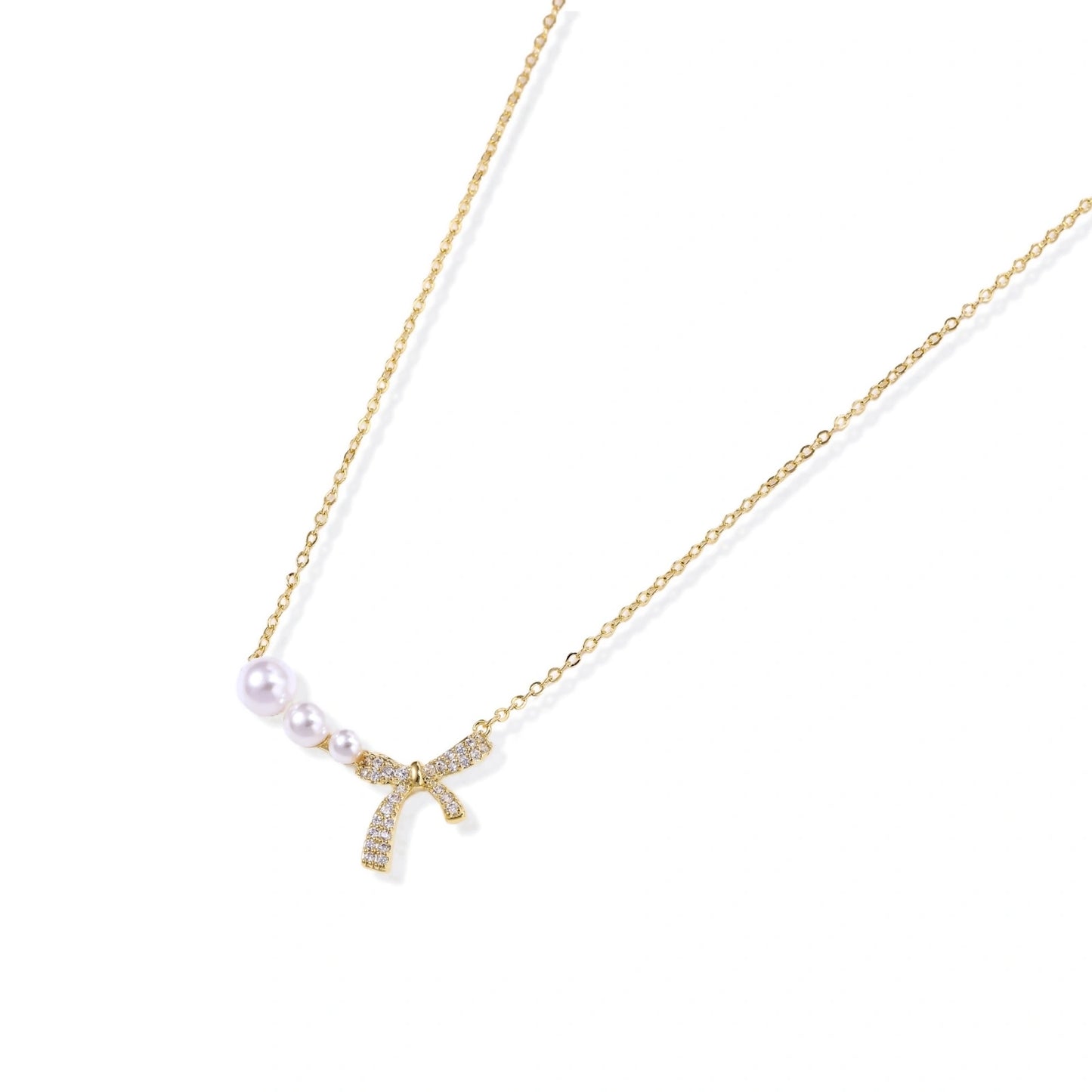 Delicate gold necklace with three white pearls and crystal-embellished bow pendant on cream background
