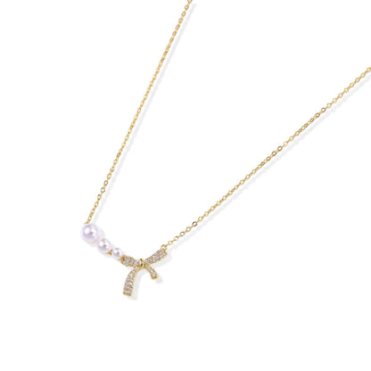 Delicate gold necklace with three white pearls and crystal-embellished bow pendant on cream background