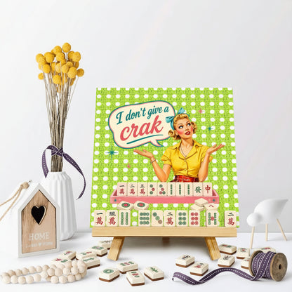 A square decorative print or canvas on a small wooden easel, featuring a retro illustration of a woman with the text 'I don't give a crac' over a green checkered background. The print is styled on a white surface with decorative props including a vase of yellow flowers, a wooden house ornament, beads, and ribbon.