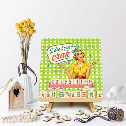 A square decorative print or canvas on a small wooden easel, featuring a retro illustration of a woman with the text 'I don't give a crac' over a green checkered background. The print is styled on a white surface with decorative props including a vase of yellow flowers, a wooden house ornament, beads, and ribbon.
