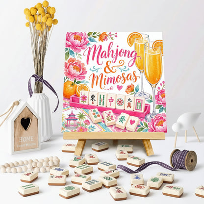 A decorative canvas print on a small wooden easel, featuring a colorful mahjong game design with the words 'Mahjong & Mimosas' and floral accents. The scene is styled on a white surface with a vase of yellow flowers, a wooden house ornament, beads, a spool of ribbon, and two glasses of orange drink.